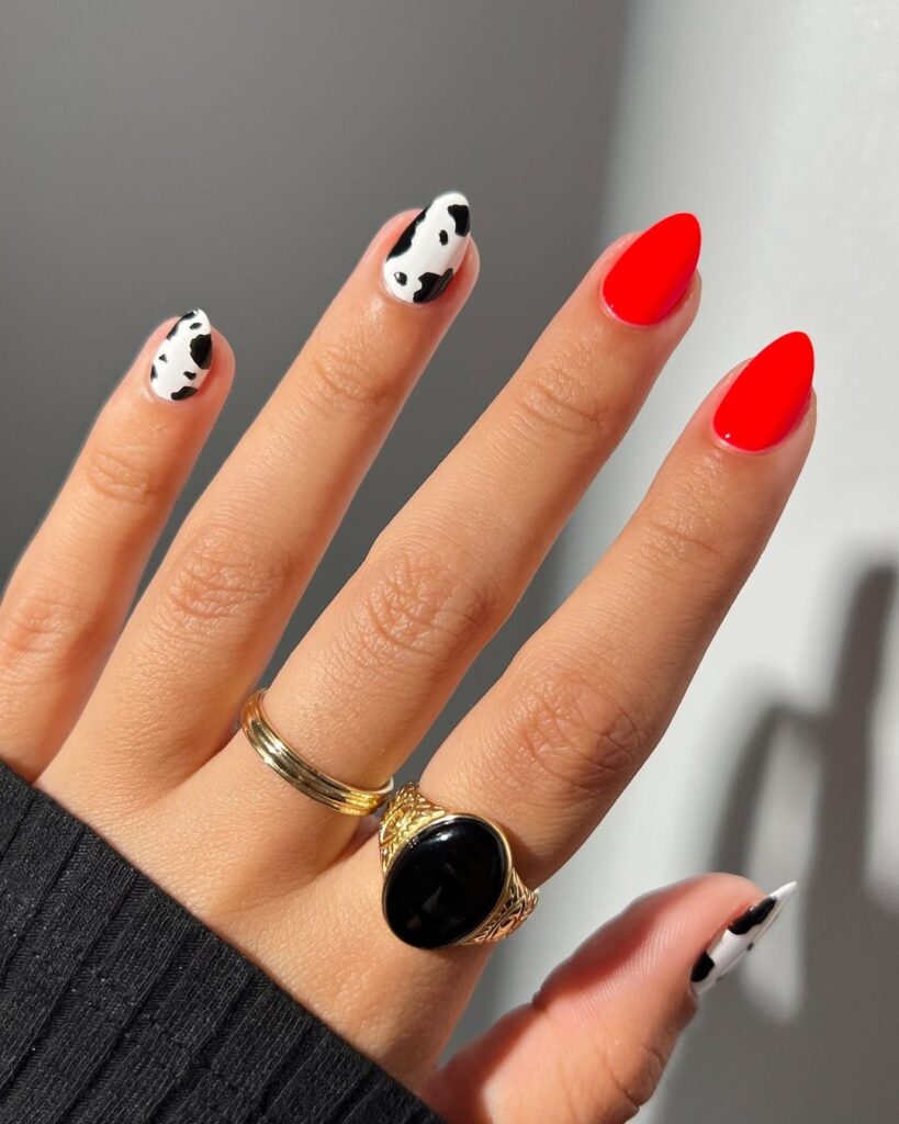 A hand with a cow print nail design on three fingers and a neon orange nail polish on two fingers.