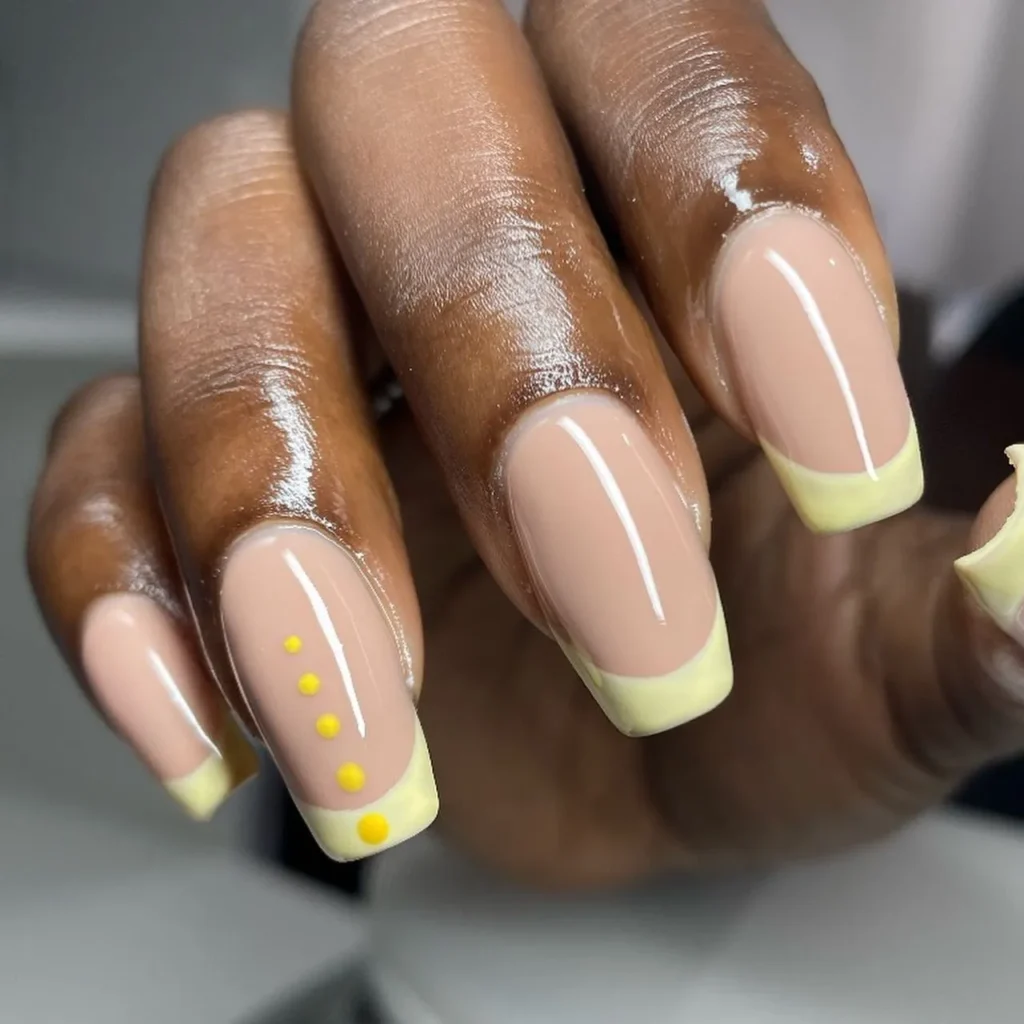 a hand with dark skin and short, square nails; painted in a light beige color with a yellow French tip and some dots on the accent nail.