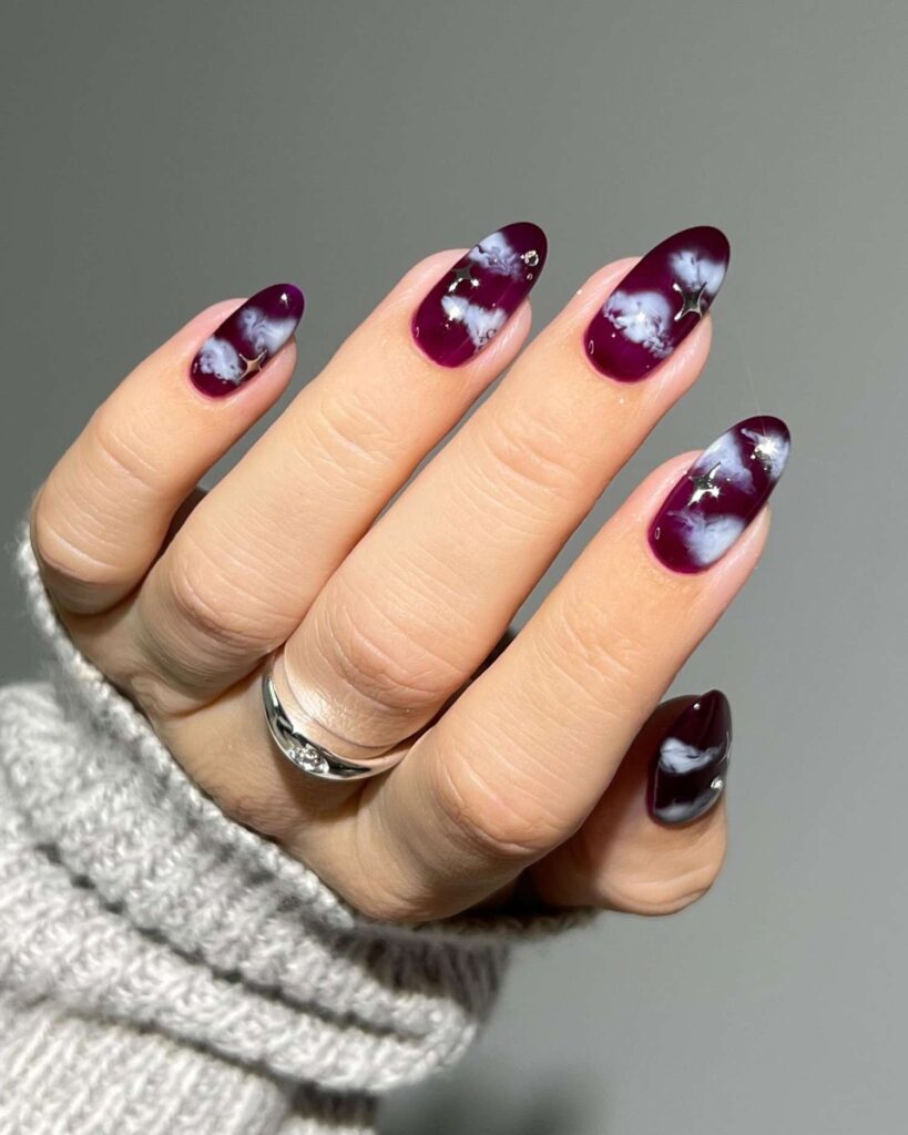 Long almond nails with a dark purple base and swirling white cloud-like patterns with silver accents